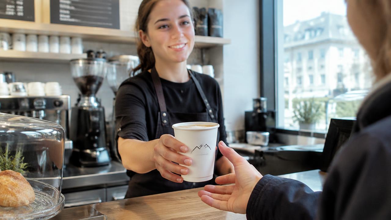 Barista reicht einen bedruckten Kaffeebecher mit Logo über die Theke